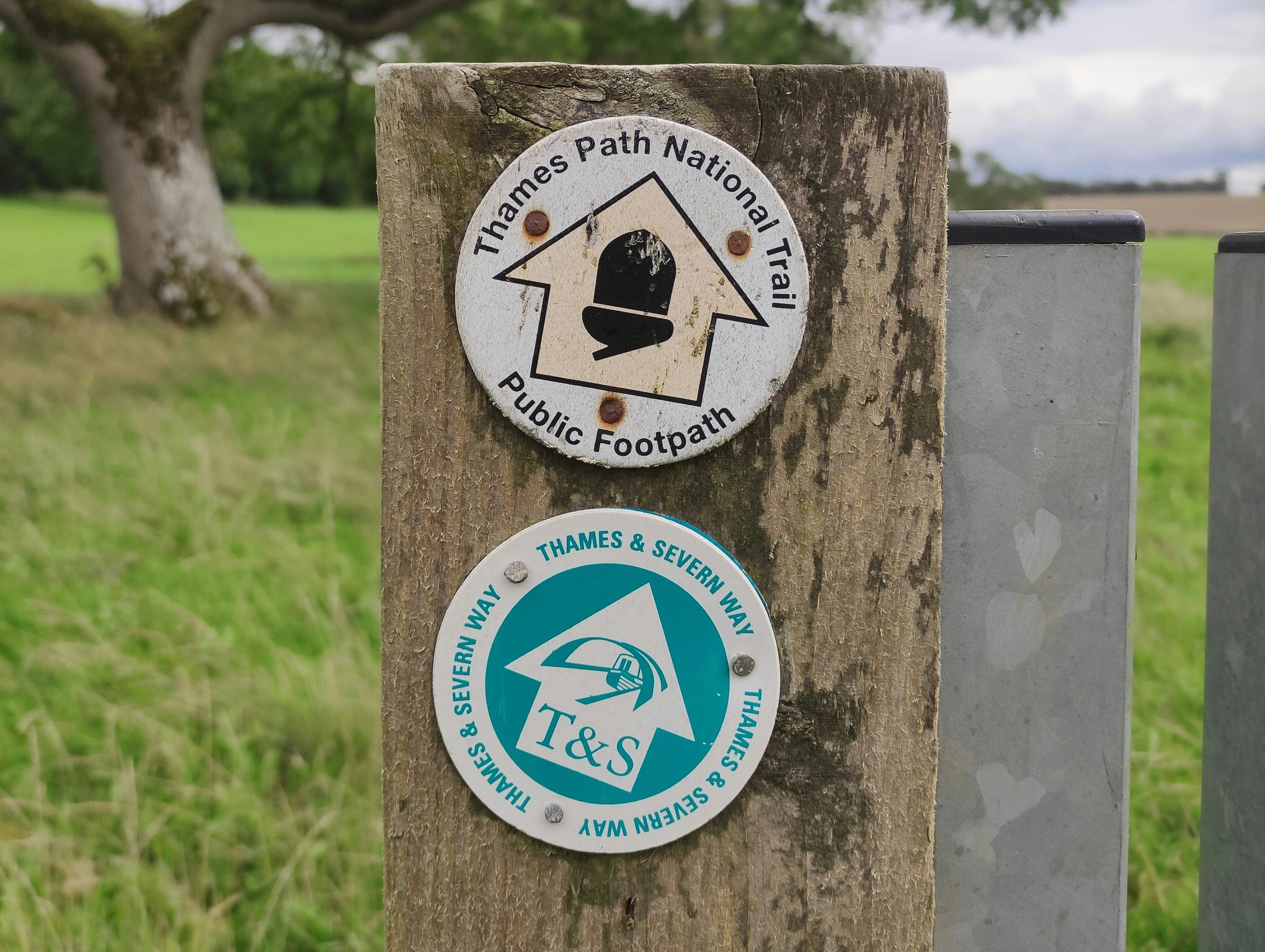 A close-up of a wooden post featuring two waymarkers for walking trails.