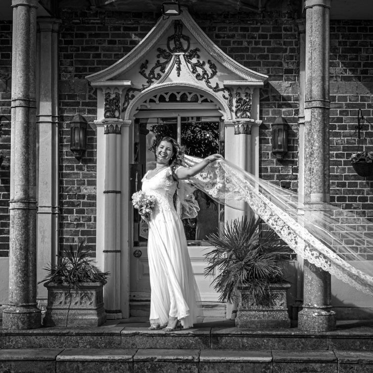 Bride stands smiling on the steps of a grand entrance, holding her bouquet as her lace veil flows dramatically in the breeze.