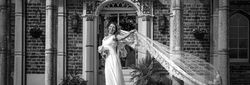 Bride stands smiling on the steps of a grand entrance, holding her bouquet as her lace veil flows dramatically in the breeze.