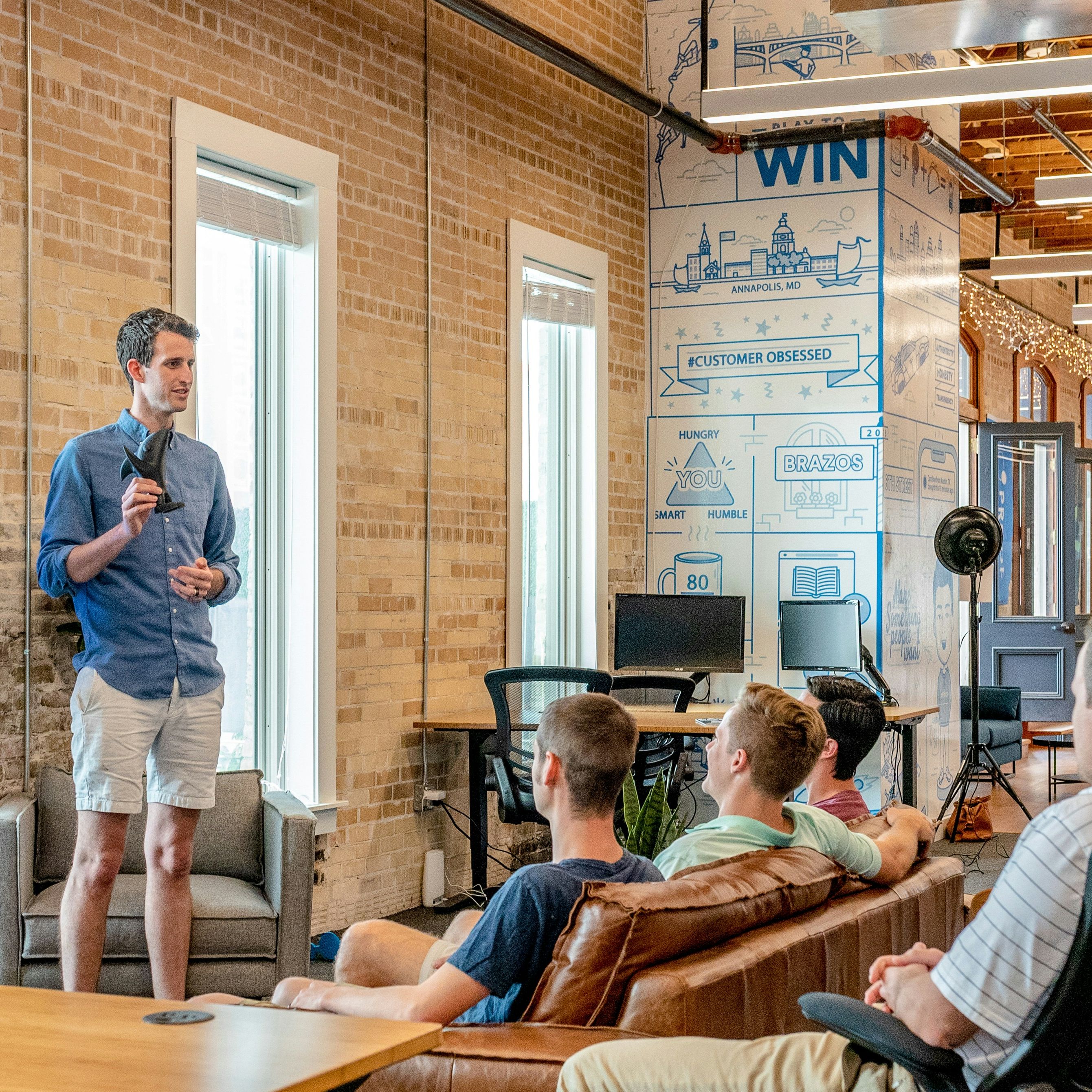 Man in shorts and a blue shirt gives a presentation to a casual office group seated on sofas, with slides displayed on a screen behind him.