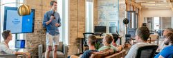 Man in shorts and a blue shirt gives a presentation to a casual office group seated on sofas, with slides displayed on a screen behind him.