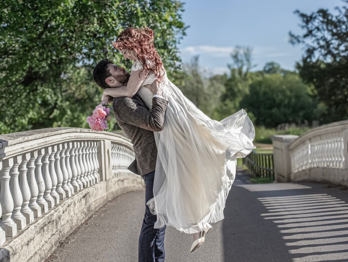 Man lifts woman in white bridal dress on a sunlit stone bridge, holding pink flowers, surrounded by greenery and trees on a clear day.