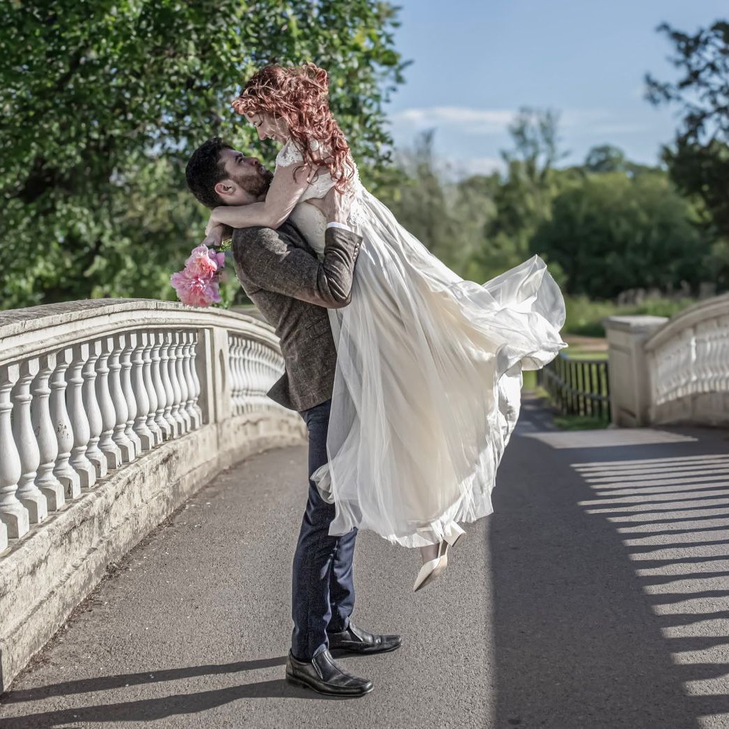 Man lifts woman in white bridal dress on a sunlit stone bridge, holding pink flowers, surrounded by greenery and trees on a clear day.