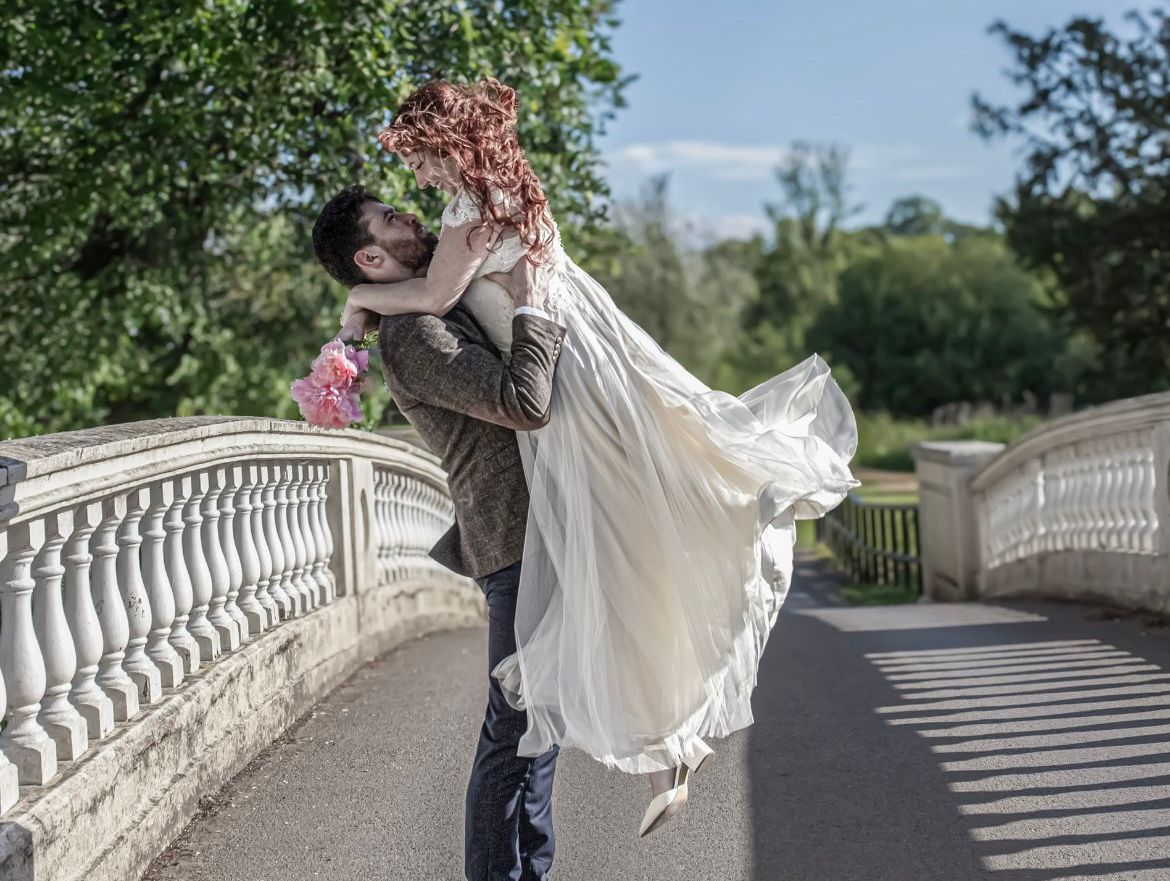 Man lifts woman in white bridal dress on a sunlit stone bridge, holding pink flowers, surrounded by greenery and trees on a clear day.