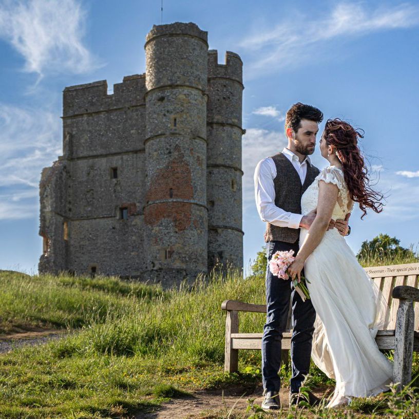 Bride and groom embrace near a wooden bench in a grassy field with an old stone castle behind them under a blue sky.