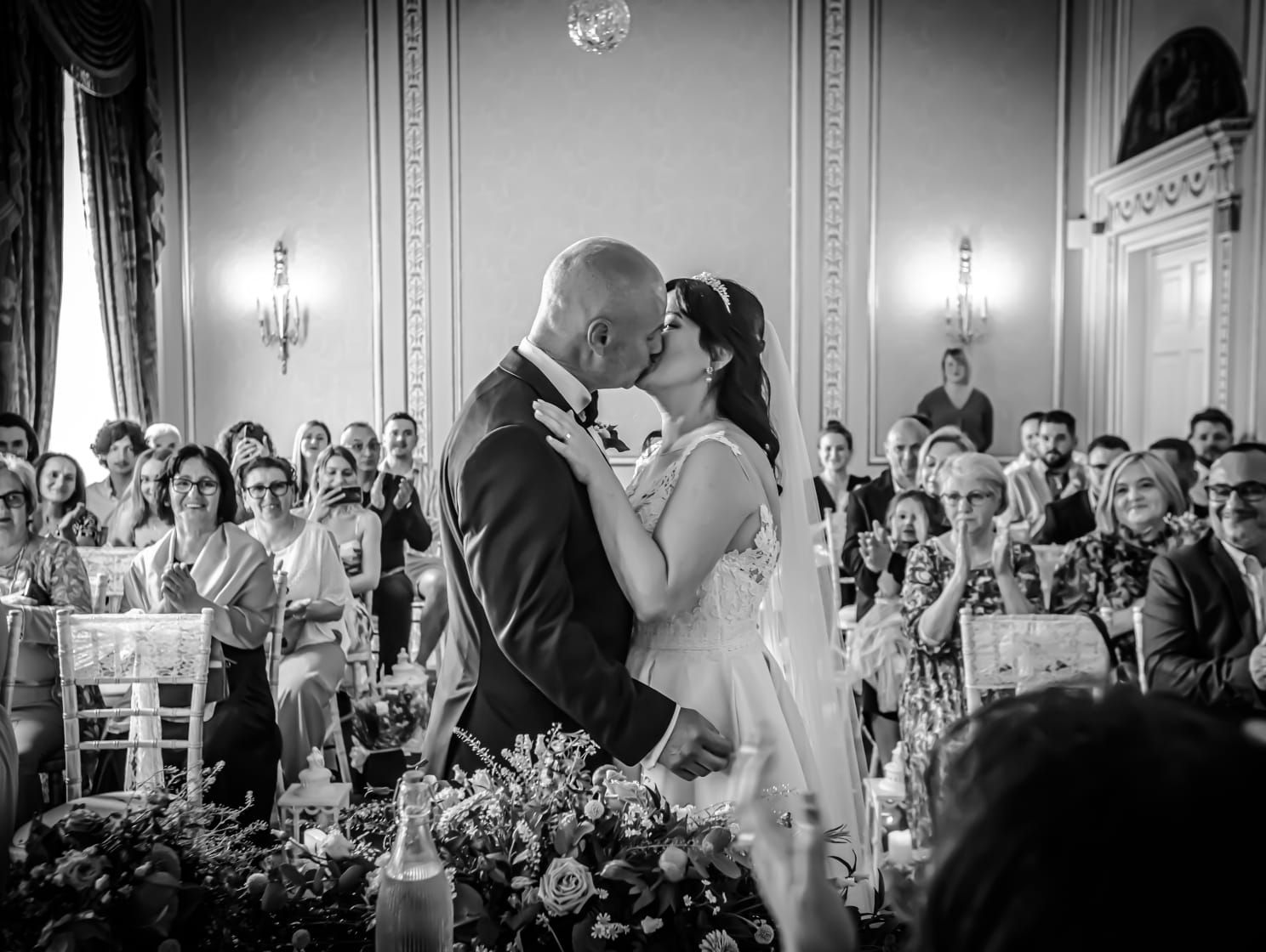 Bride and groom share a kiss during their wedding ceremony, surrounded by smiling guests clapping in a grand, elegant room.