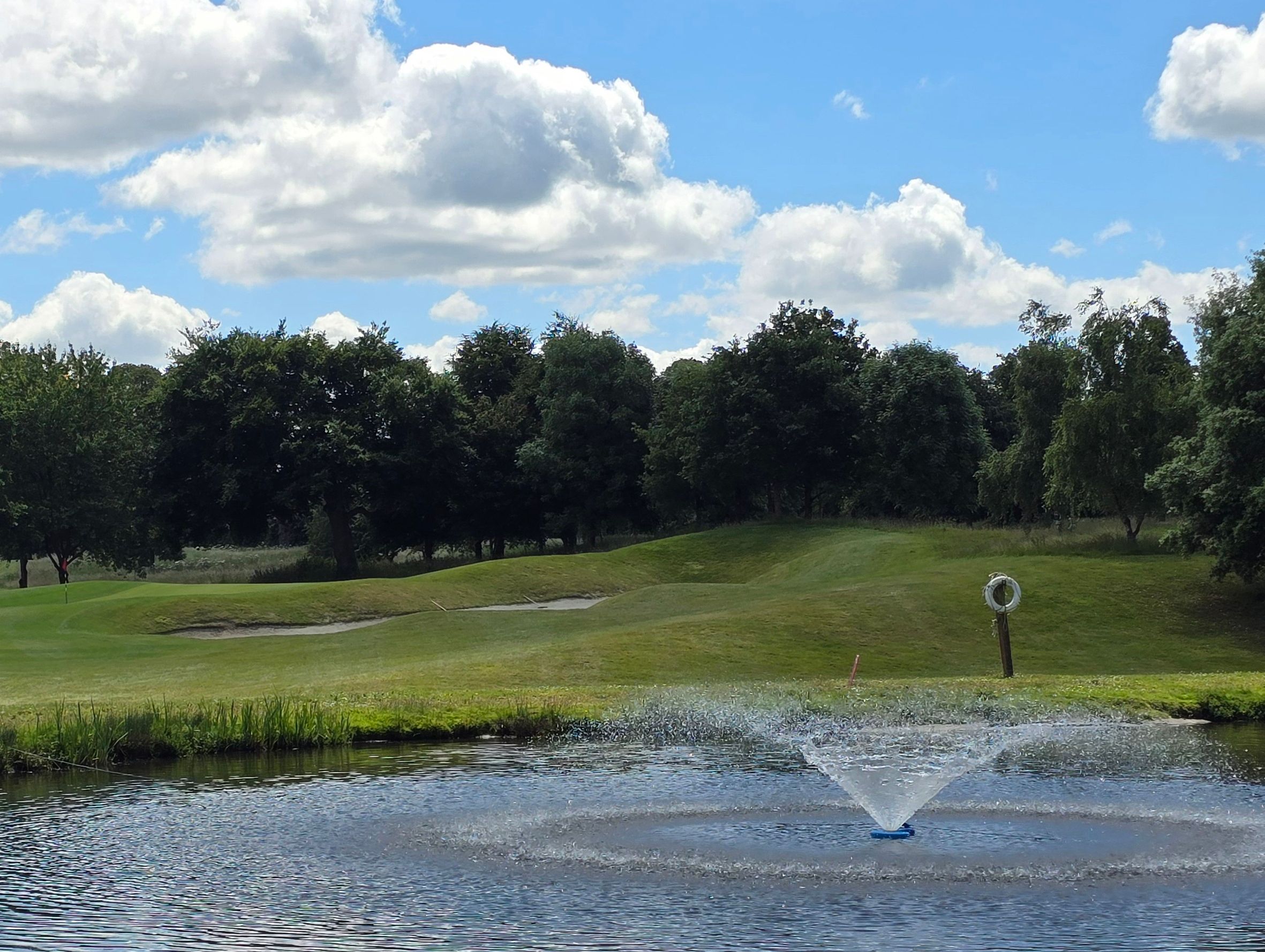Golf course with a pond and water fountain in the foreground, surrounded by trees and a blue sky with clouds.