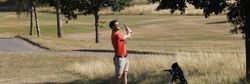Man playing golf on a sunny day near a tree, swinging a golf club with a golf bag nearby.