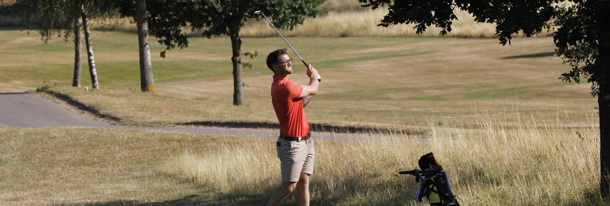 Man playing golf on a sunny day near a tree, swinging a golf club with a golf bag nearby.