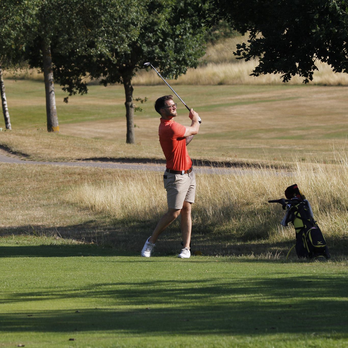 Man playing golf on a sunny day near a tree, swinging a golf club with a golf bag nearby.
