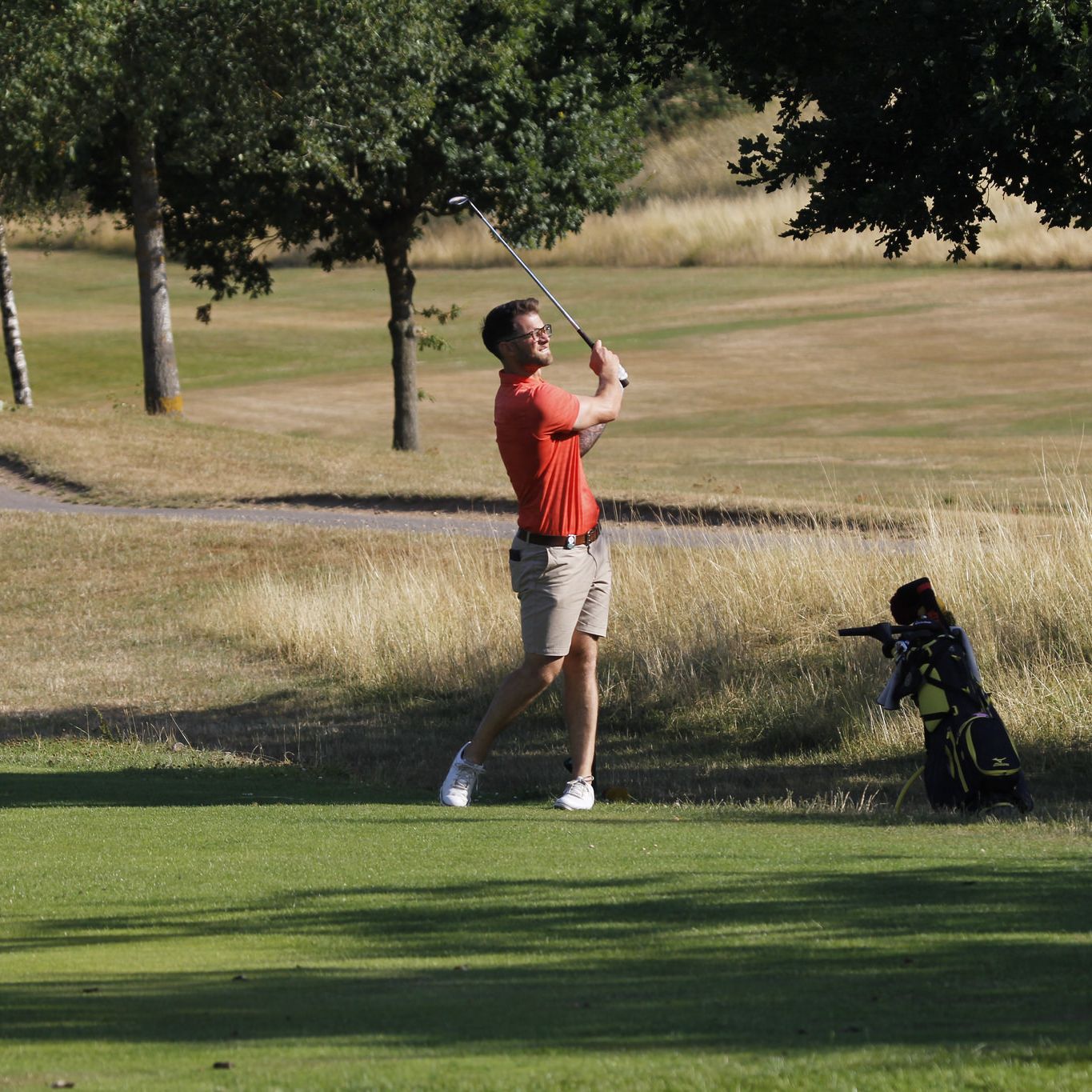Man playing golf on a sunny day near a tree, swinging a golf club with a golf bag nearby.