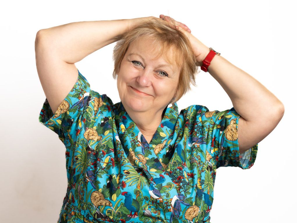 Smiling woman with short blonde hair holding her hands on top of her head, wearing a colorful printed shirt