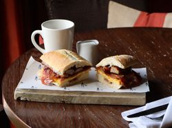 A breakfast sandwich with sausage, egg and bacon on a ciabatta roll with a coffee cup on a dark wood table