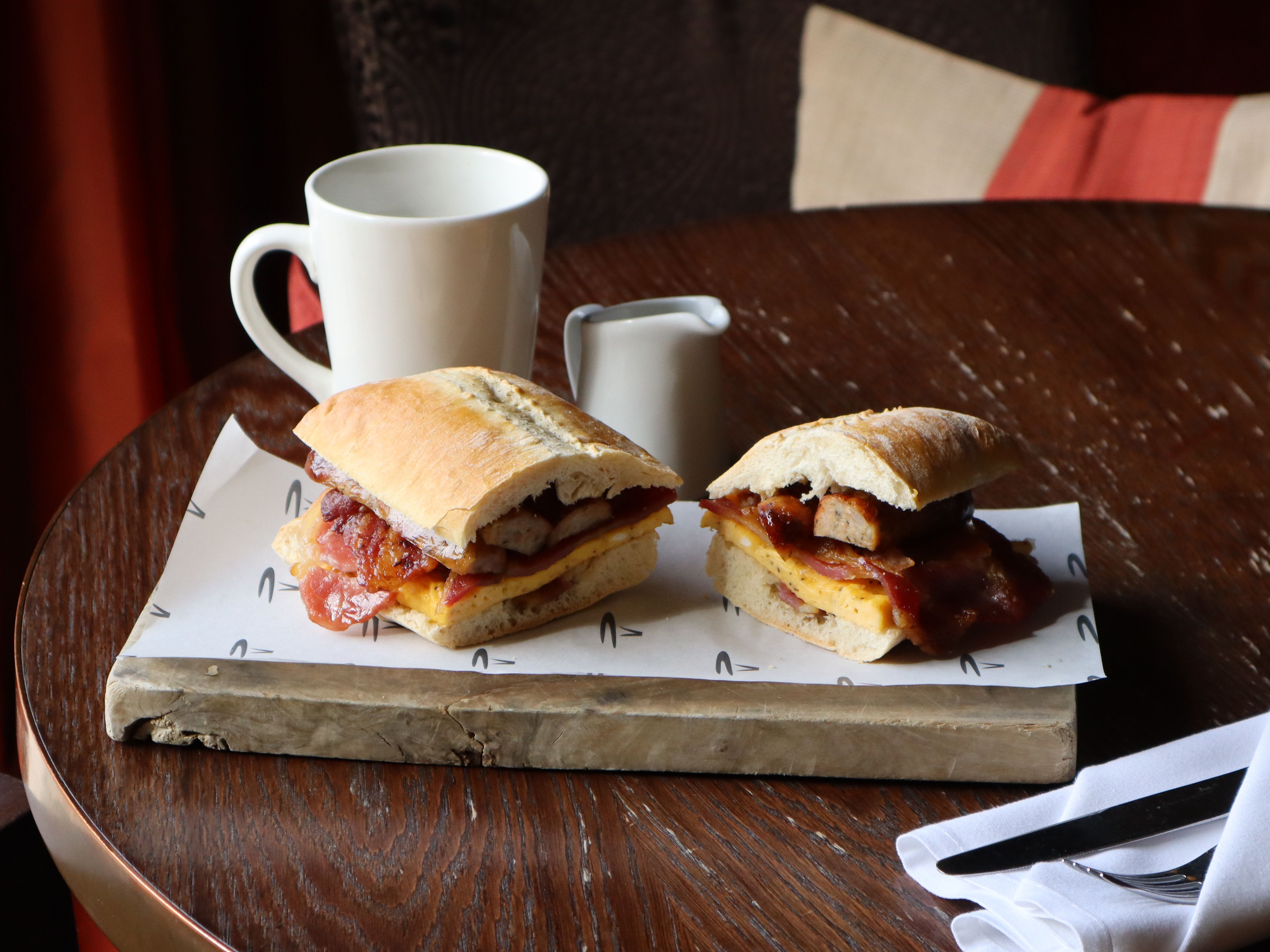 A breakfast sandwich with sausage, egg and bacon on a ciabatta roll with a coffee cup on a dark wood table
