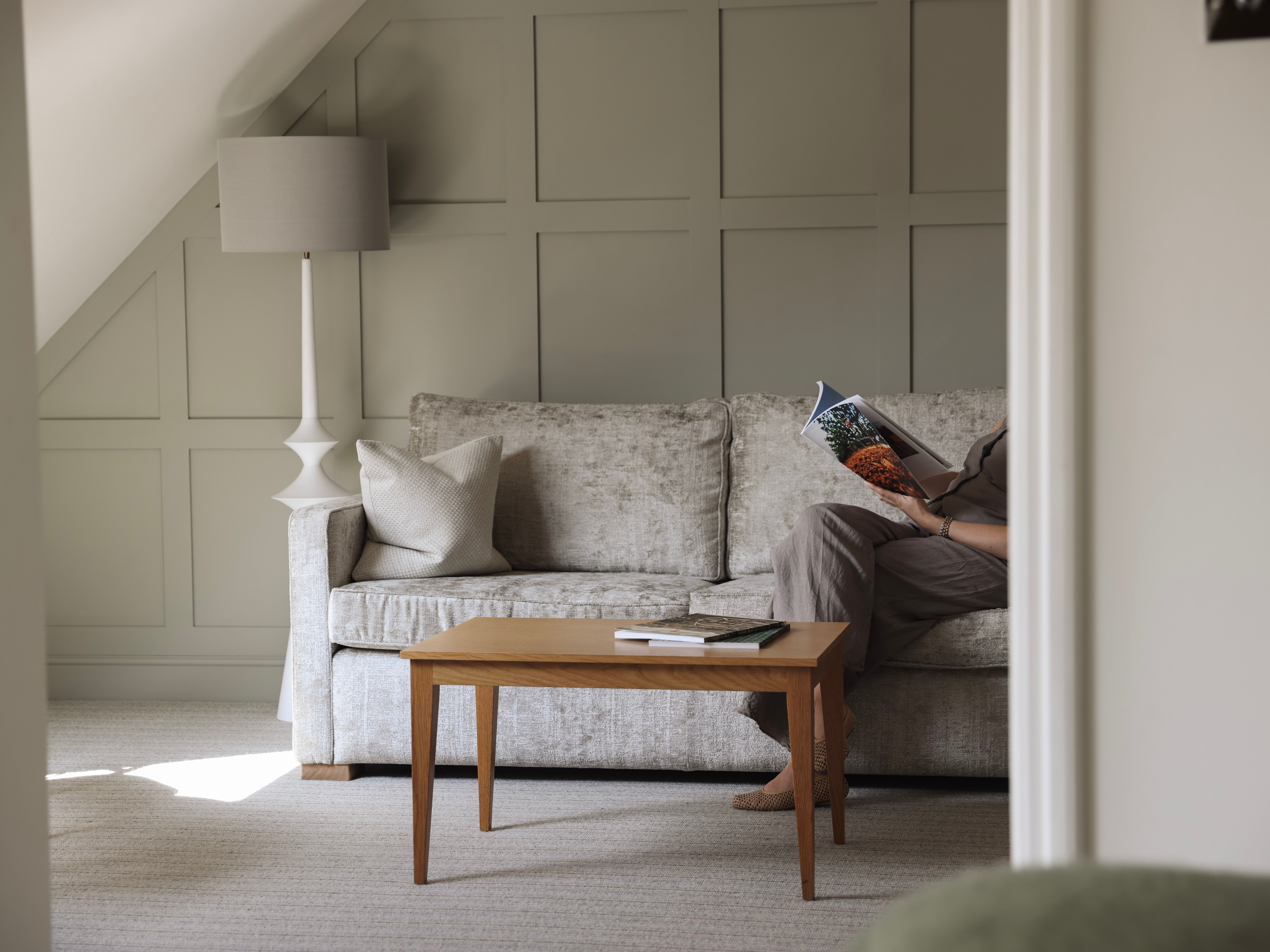 A cozy living area with a light grey sofa, wooden coffee table, and a person reading a magazine.