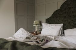 A cozy bed with white linens, a green blanket, and a newspaper lying open. A lamp and mug sit on the bedside table.