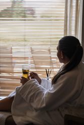 Person in a white robe relaxing with a cup of tea by a window with blinds