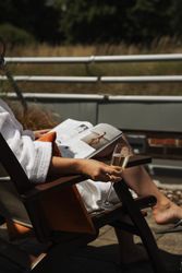 Person in a white robe relaxing on a chair outdoors, holding a glass of champagne and reading a magazine.