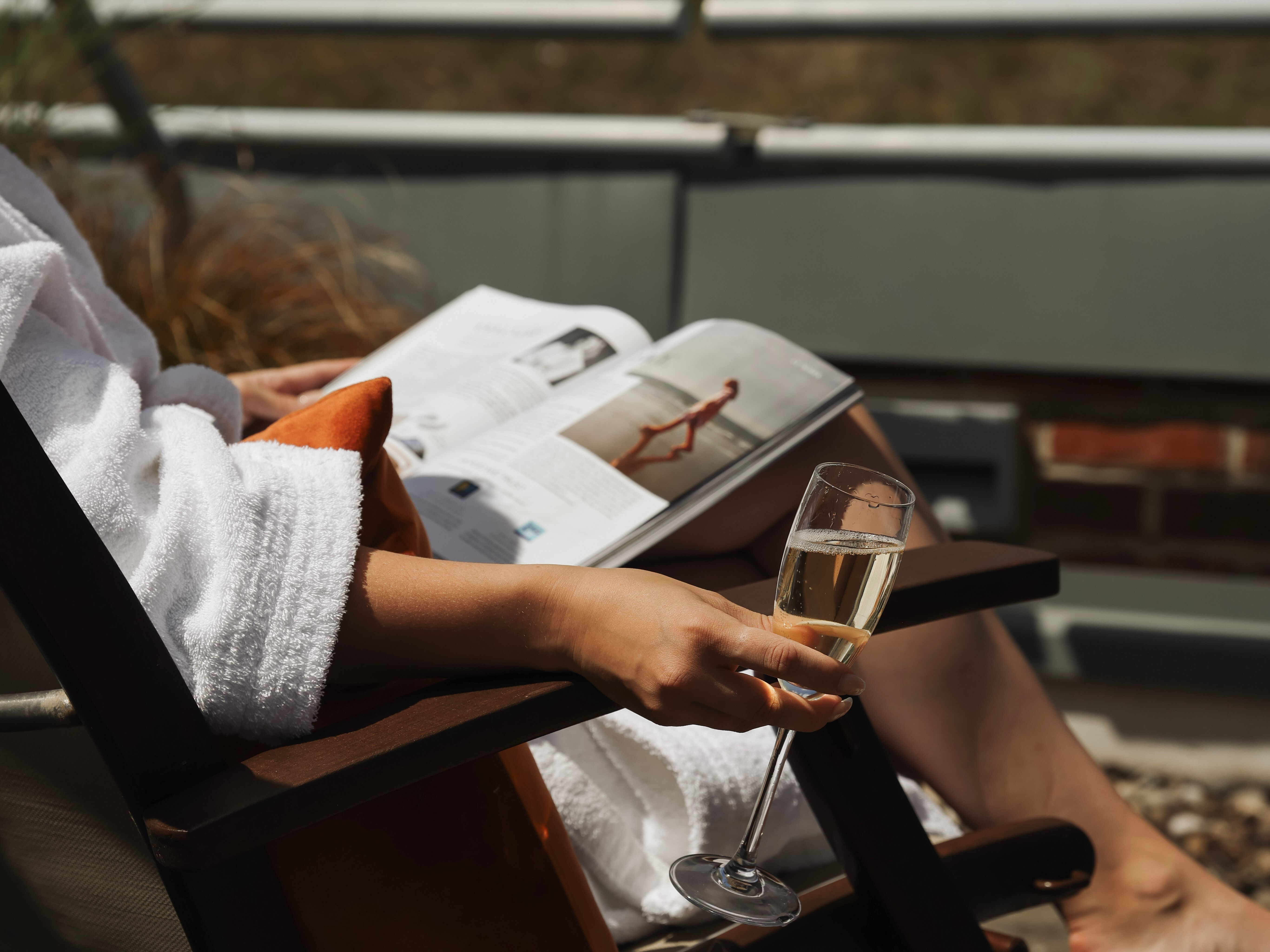 Person in a white robe relaxing on a chair outdoors, holding a glass of champagne and reading a magazine.