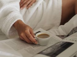 Person in a white bathrobe holding a cup of coffee while reading a magazine in bed