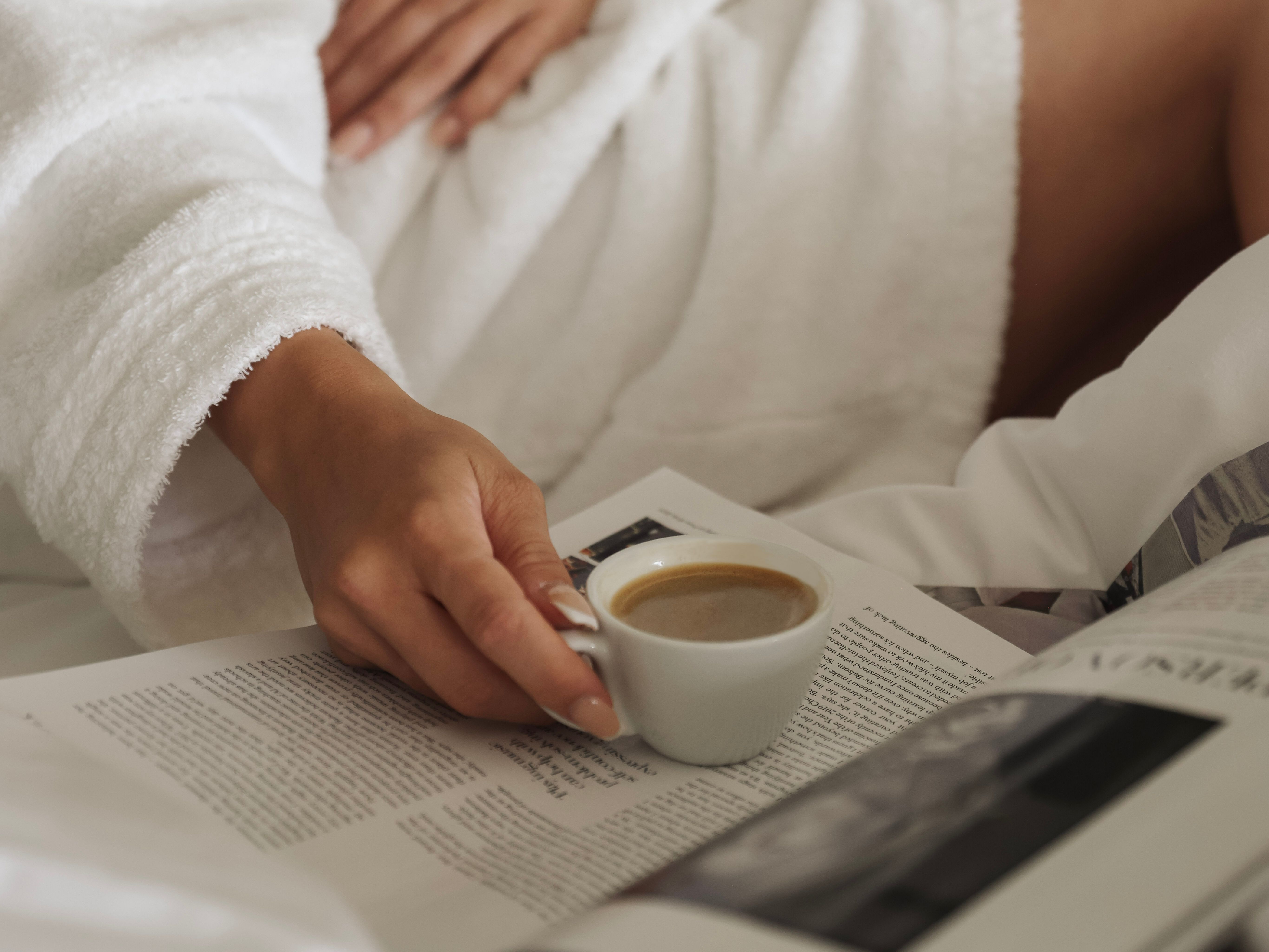 Person in a white bathrobe holding a cup of coffee while reading a magazine in bed