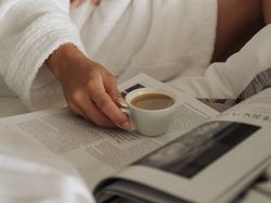 Person in a white bathrobe holding a cup of coffee while reading a magazine in bed