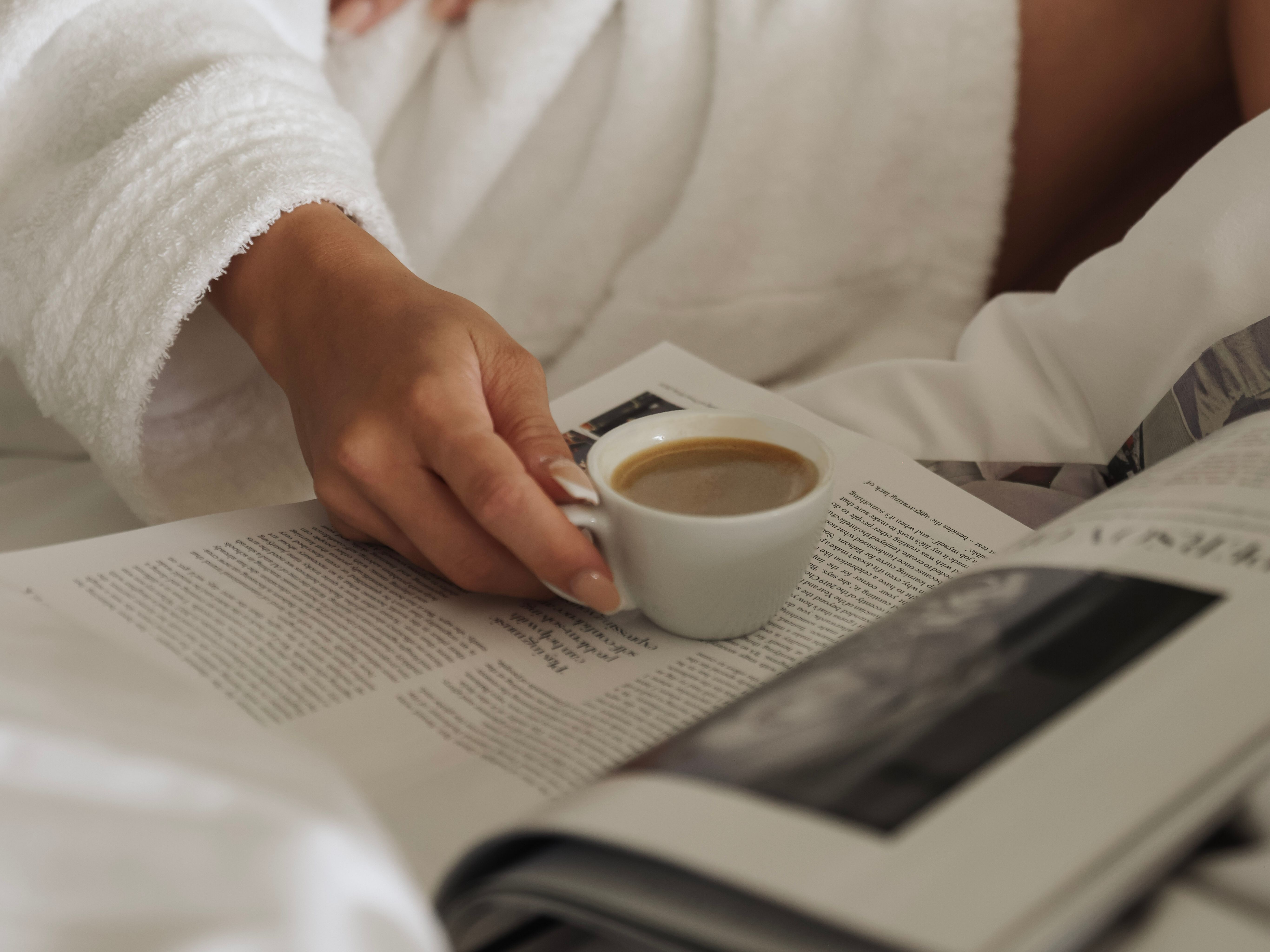 Person in a white bathrobe holding a cup of coffee while reading a magazine in bed