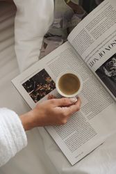 A person holding a cup of coffee over an open magazine while sitting on a bed.