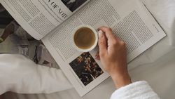 A person in a white robe holding a cup of coffee over an open magazine in bed