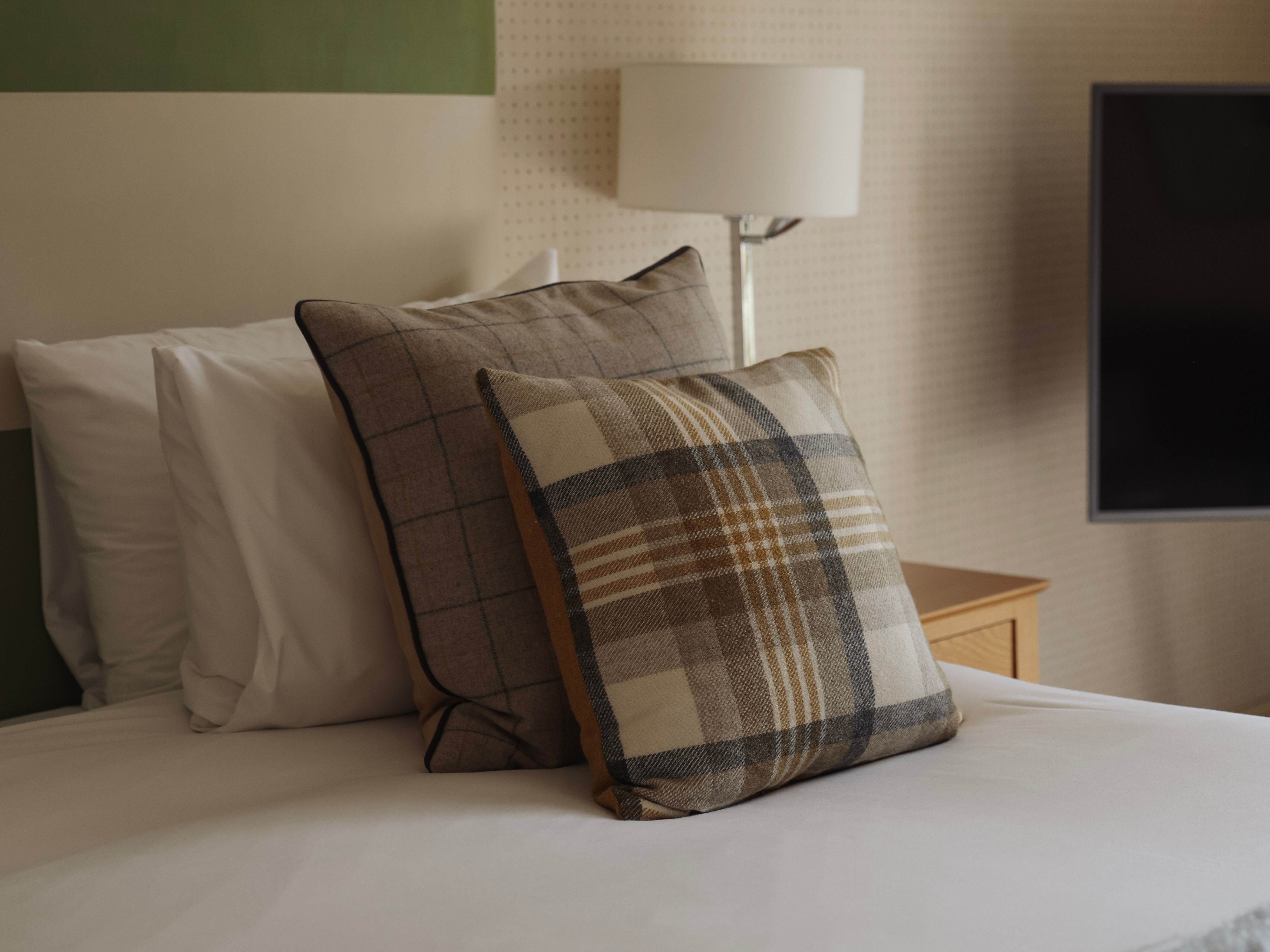 Neatly made bed with plaid decorative pillows, a green and beige headboard, and a bedside lamp.