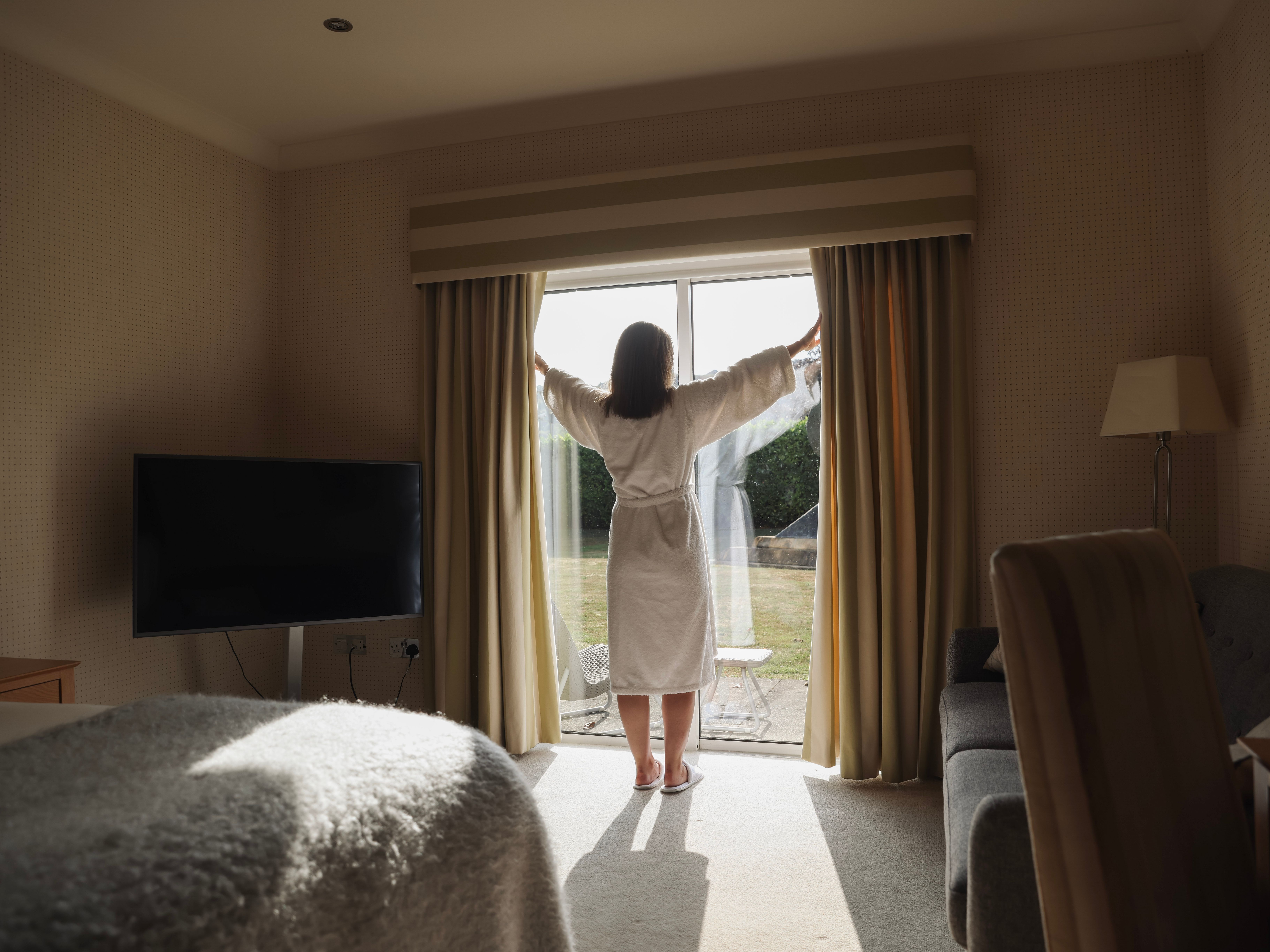 Person in a bathrobe opening curtains in a sunlit hotel room
