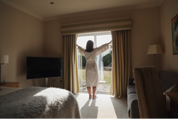 Person in a bathrobe opening curtains in a sunlit hotel room
