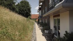 Modern building with outdoor seating along a paved path and tall grass on one side.