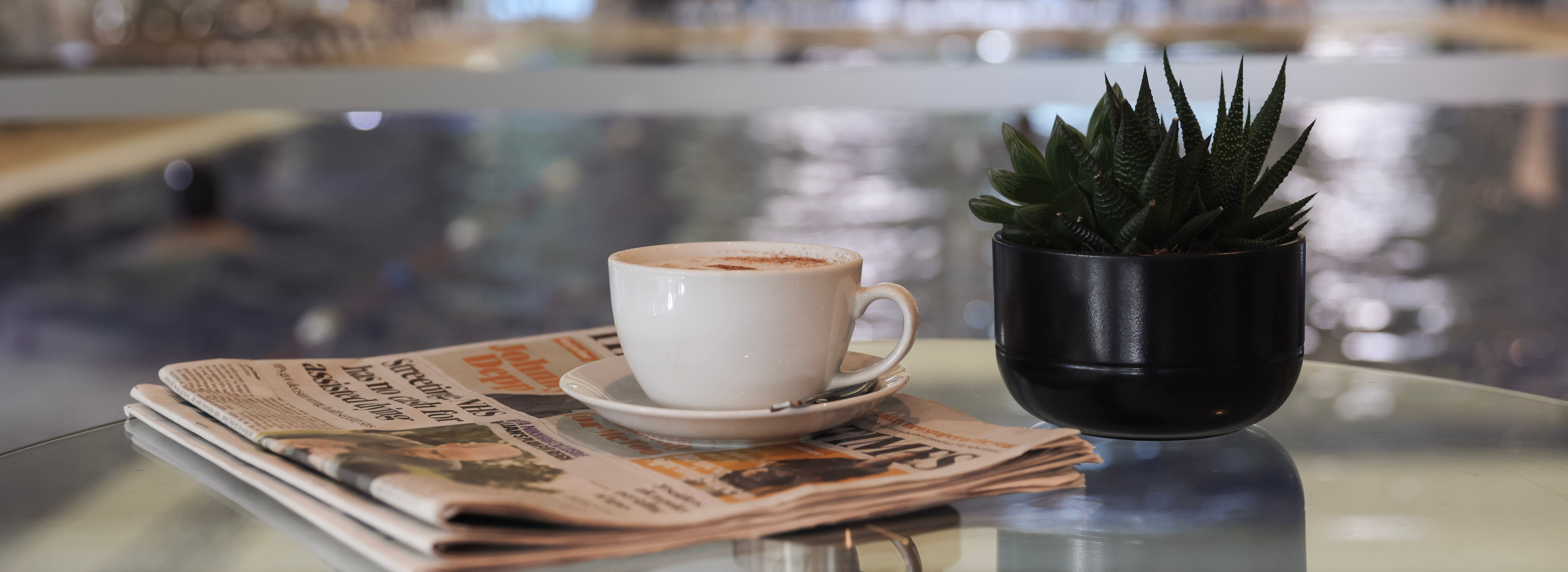 A cup of coffee, a small potted succulent, and a newspaper on a glass table with a blurred indoor pool in the background.