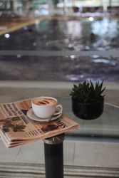 A cup of cappuccino on a newspaper next to a small potted succulent on a glass table by an indoor swimming pool.