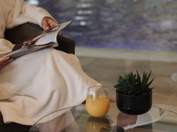 Person in a white robe reading a magazine by an indoor pool with a glass of orange juice and a potted succulent on a glass table.