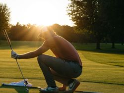 Golfer squatting on the green at sunset, preparing for a putt.