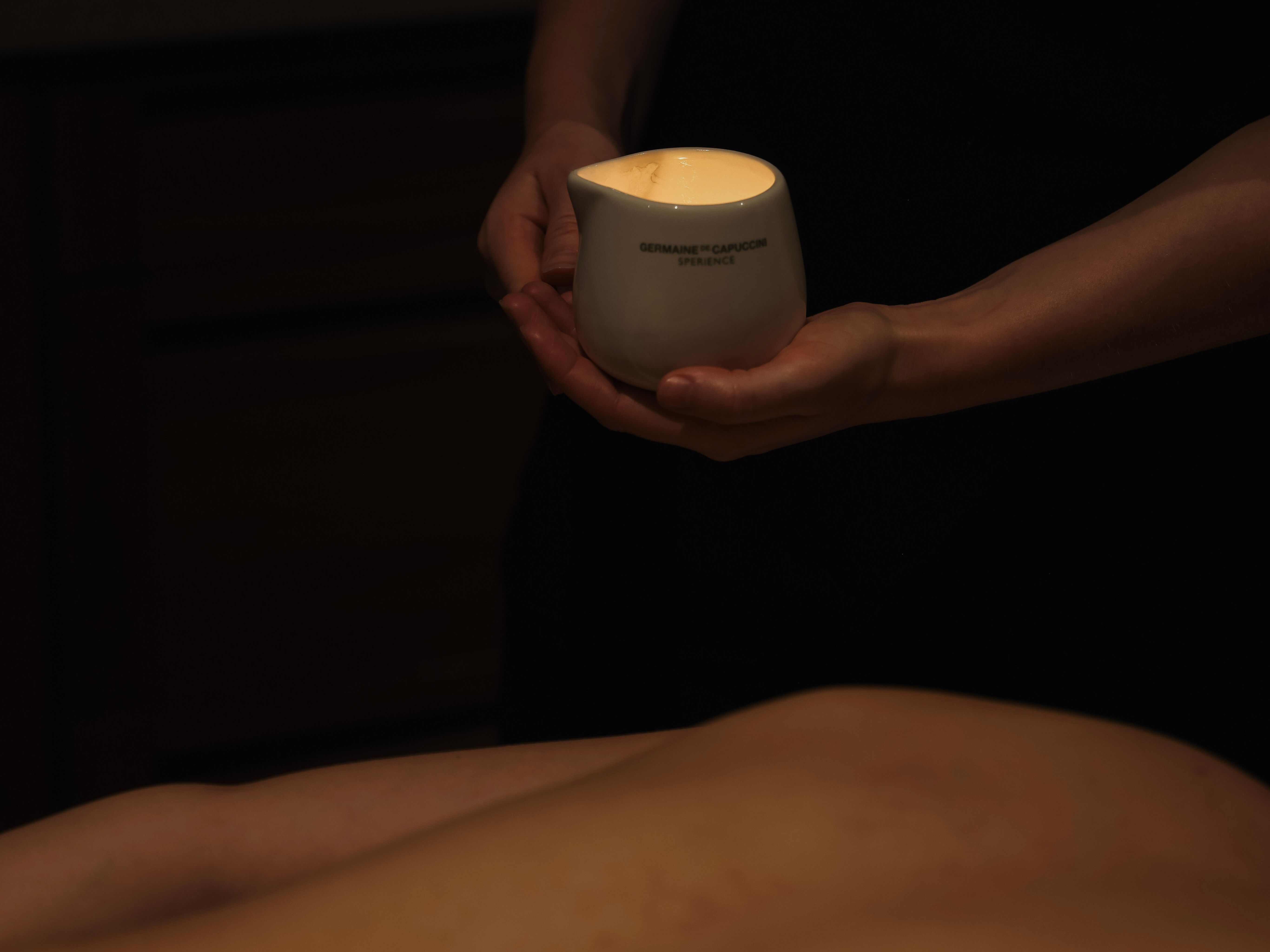 A person holds a white ceramic container with a lit candle inside during a massage session.