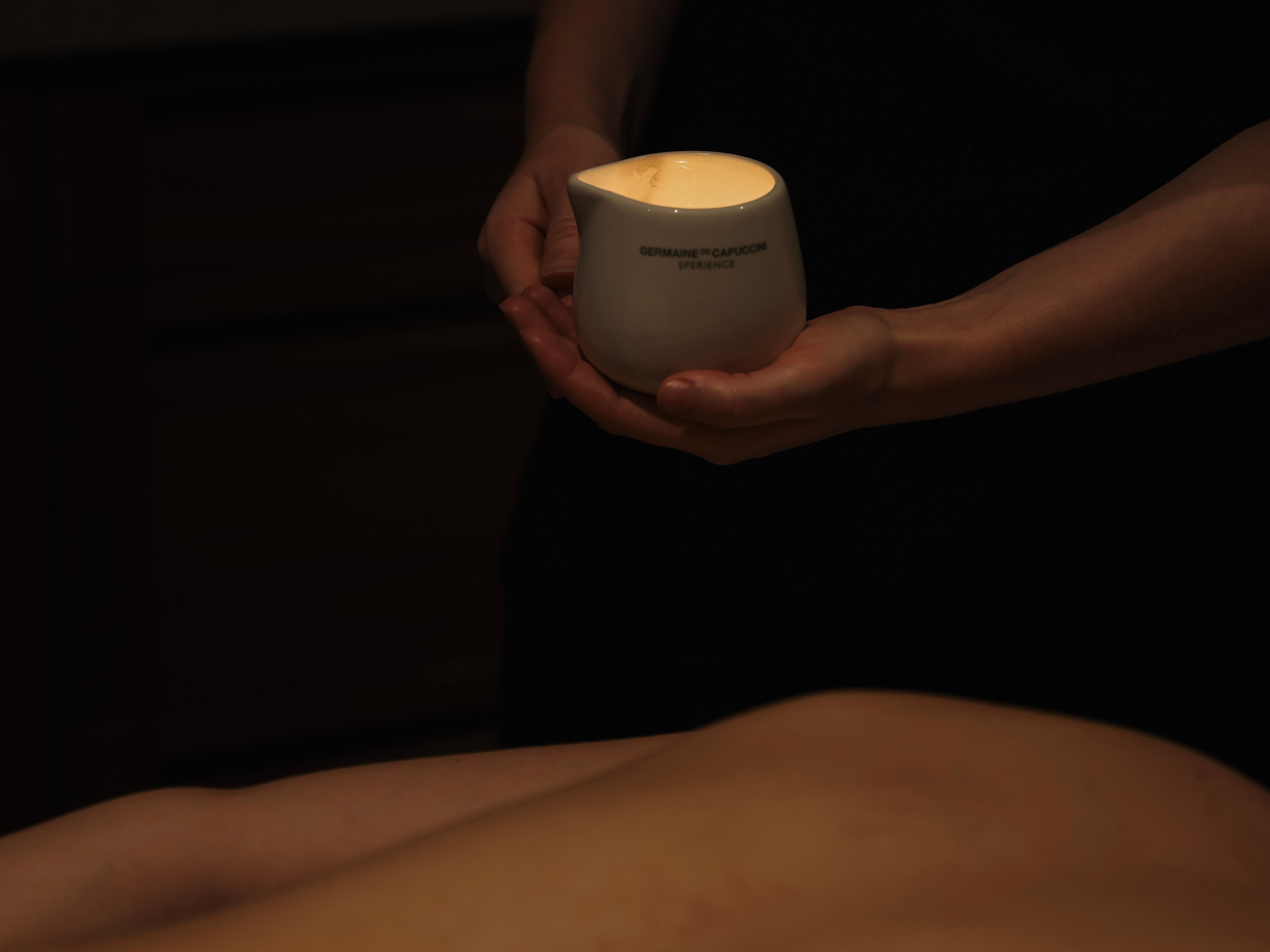 A person holds a white ceramic container with a lit candle inside during a massage session.