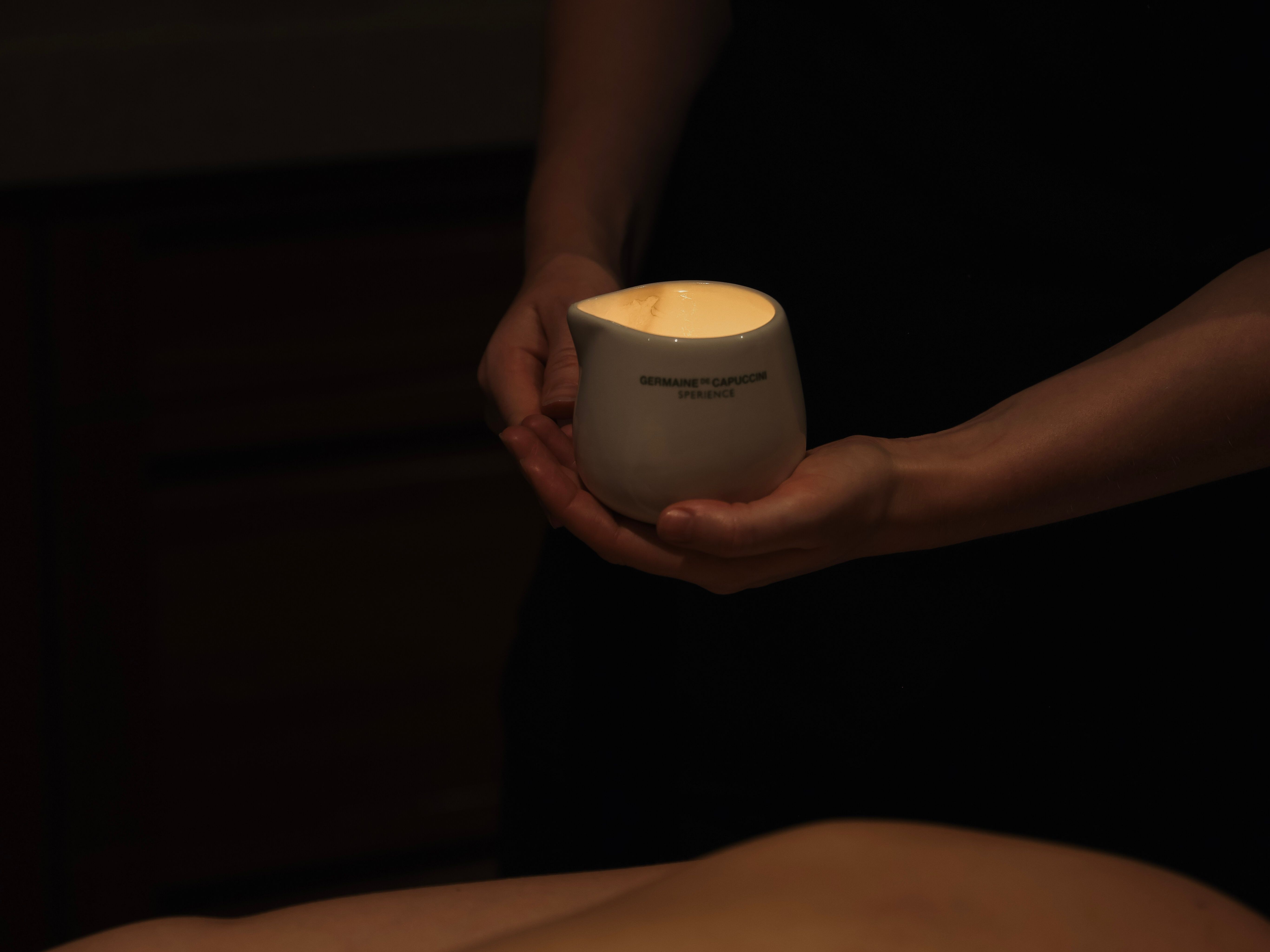A person holds a white ceramic container with a lit candle inside during a massage session.