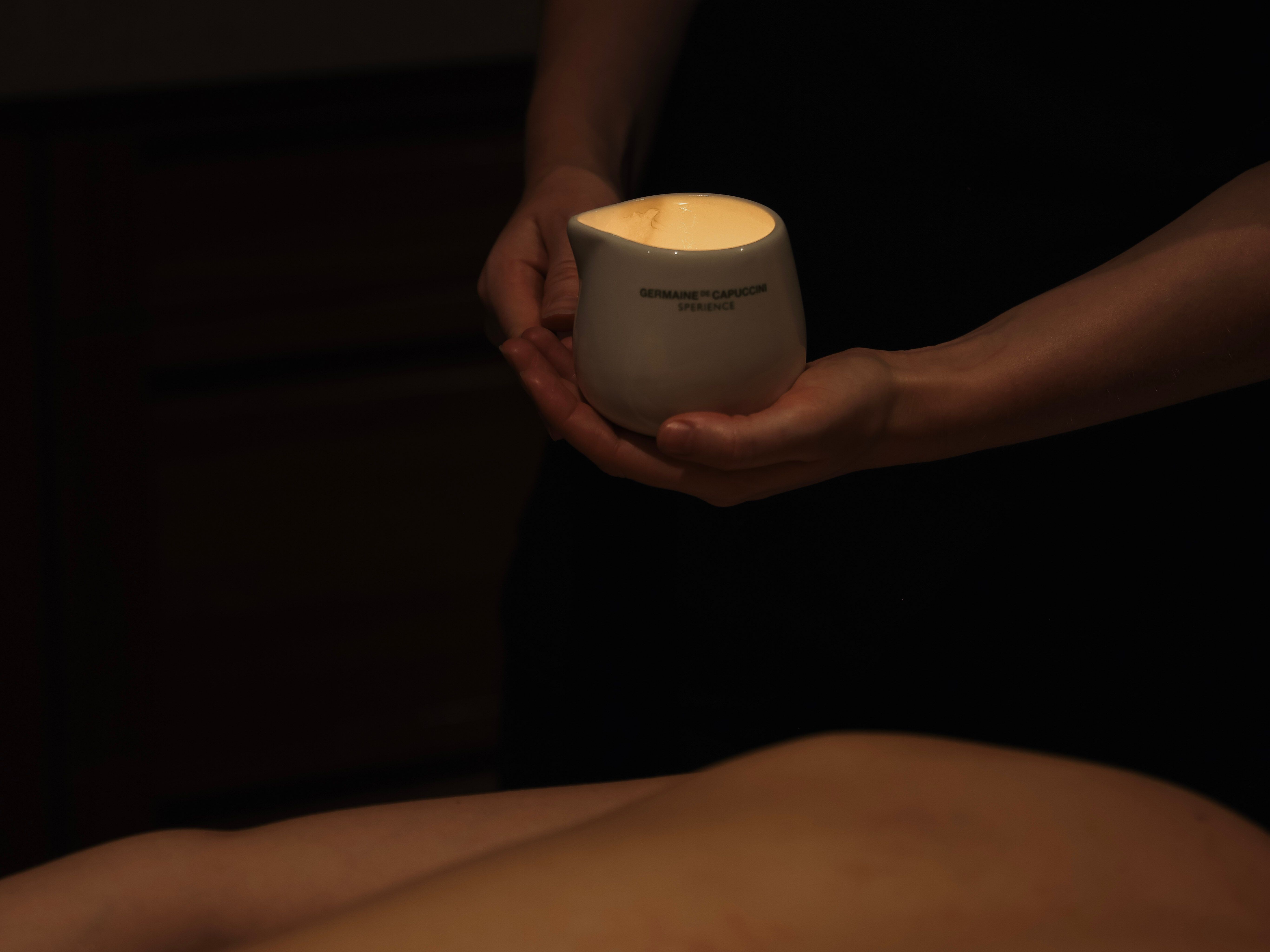 A person holds a white ceramic container with a lit candle inside during a massage session.