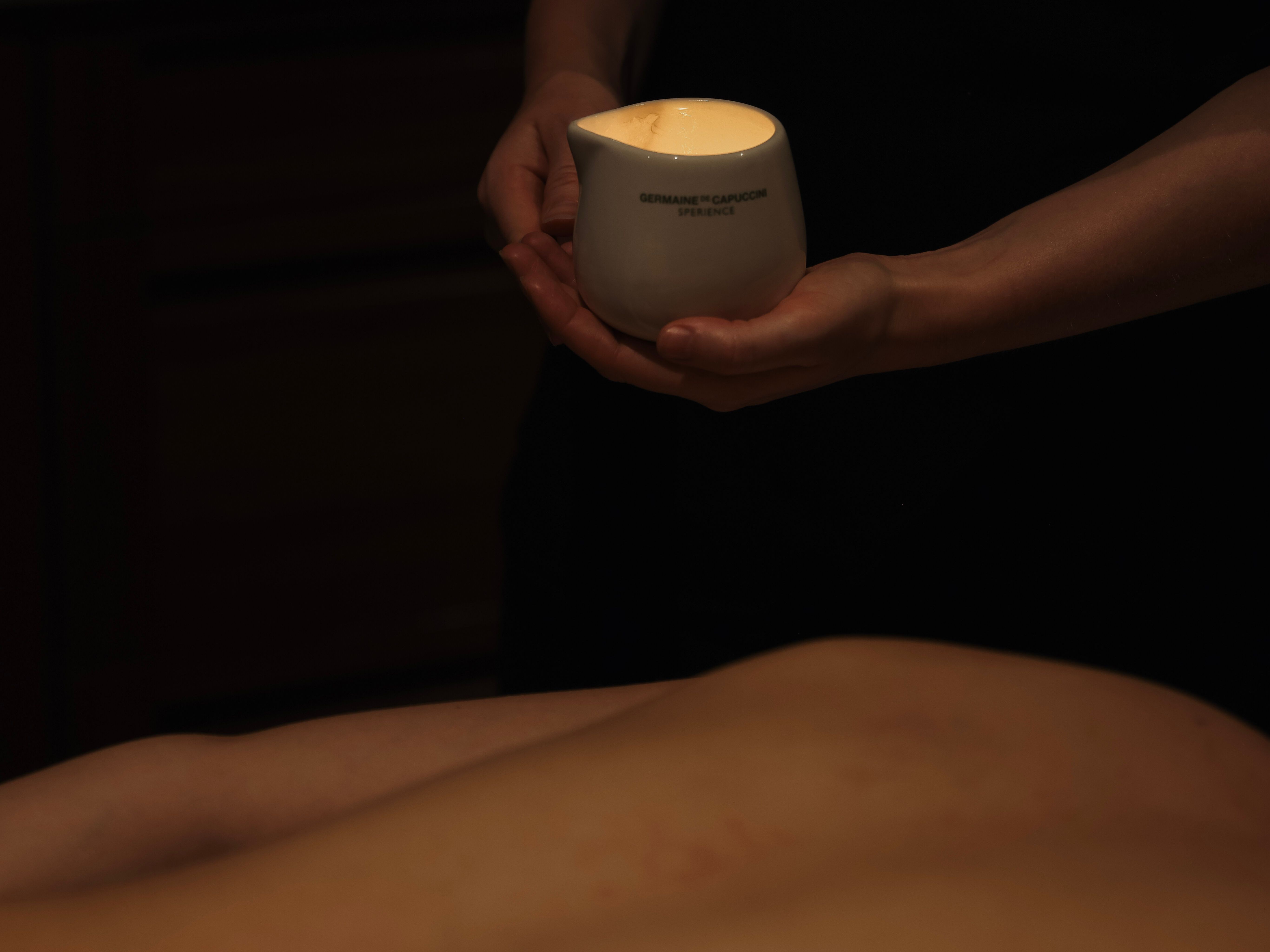 A person holds a white ceramic container with a lit candle inside during a massage session.