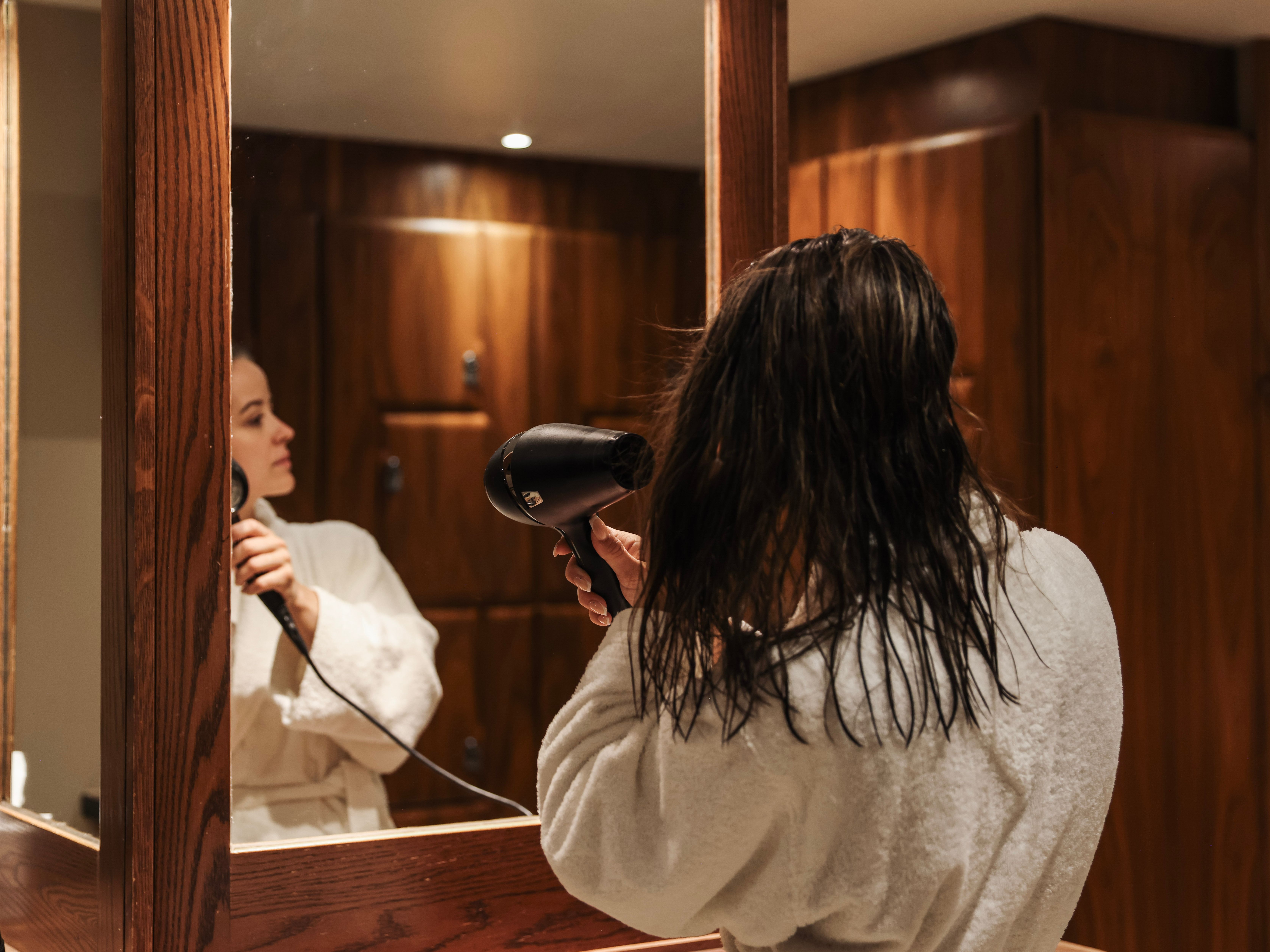 Person in a white bathrobe blow drying their wet hair in front of a mirror in a wooden-paneled bathroom.