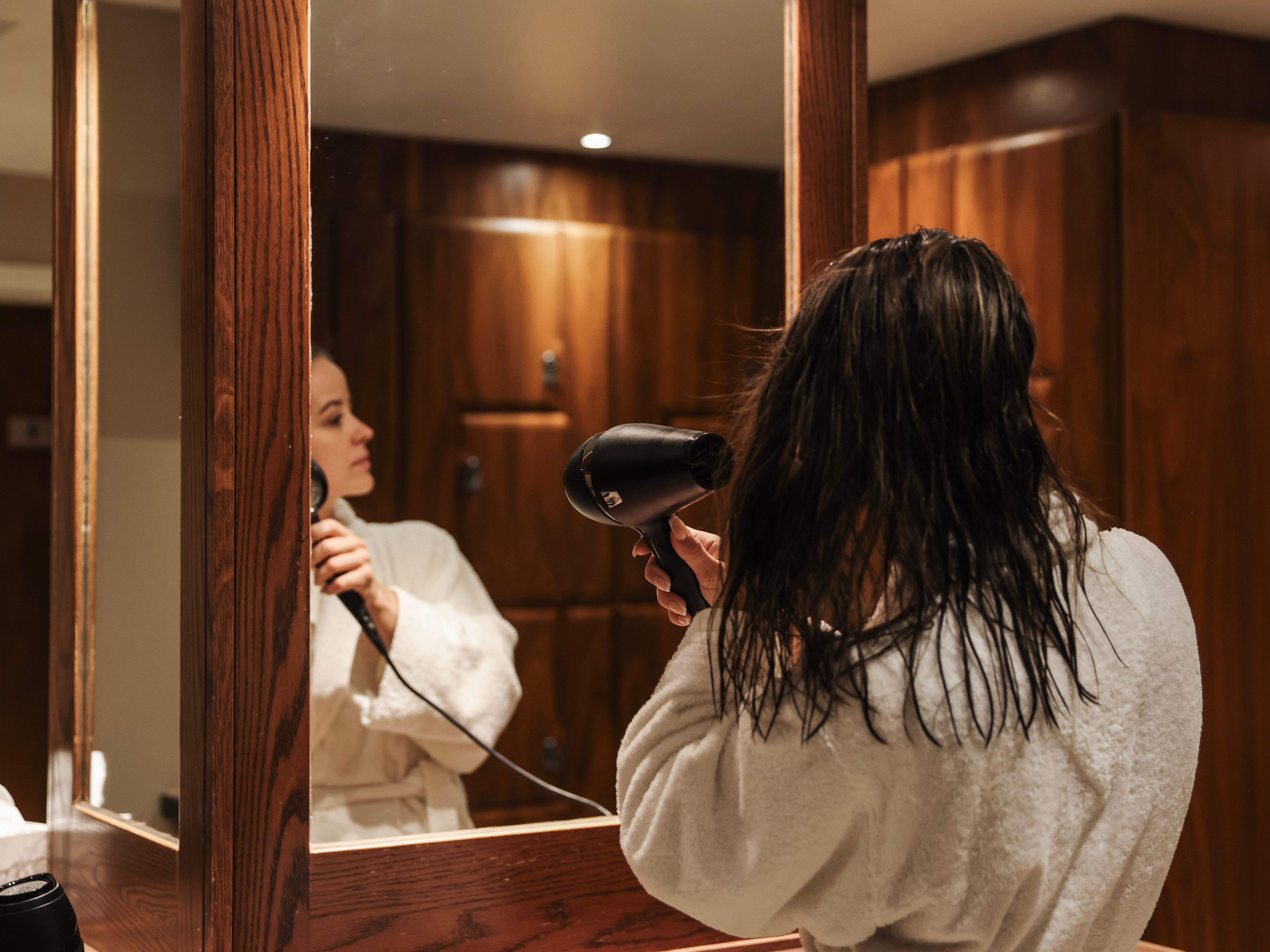 Person in a white bathrobe blow drying their wet hair in front of a mirror in a wooden-paneled bathroom.