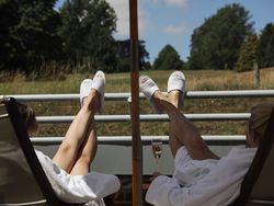 Two people relaxing on lounge chairs in white robes and slippers, with their legs up on a balcony railing, enjoying a view of a grassy field and trees