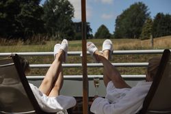 Two people relaxing on lounge chairs in white robes and slippers, with their legs up on a balcony railing, enjoying a view of a grassy field and trees