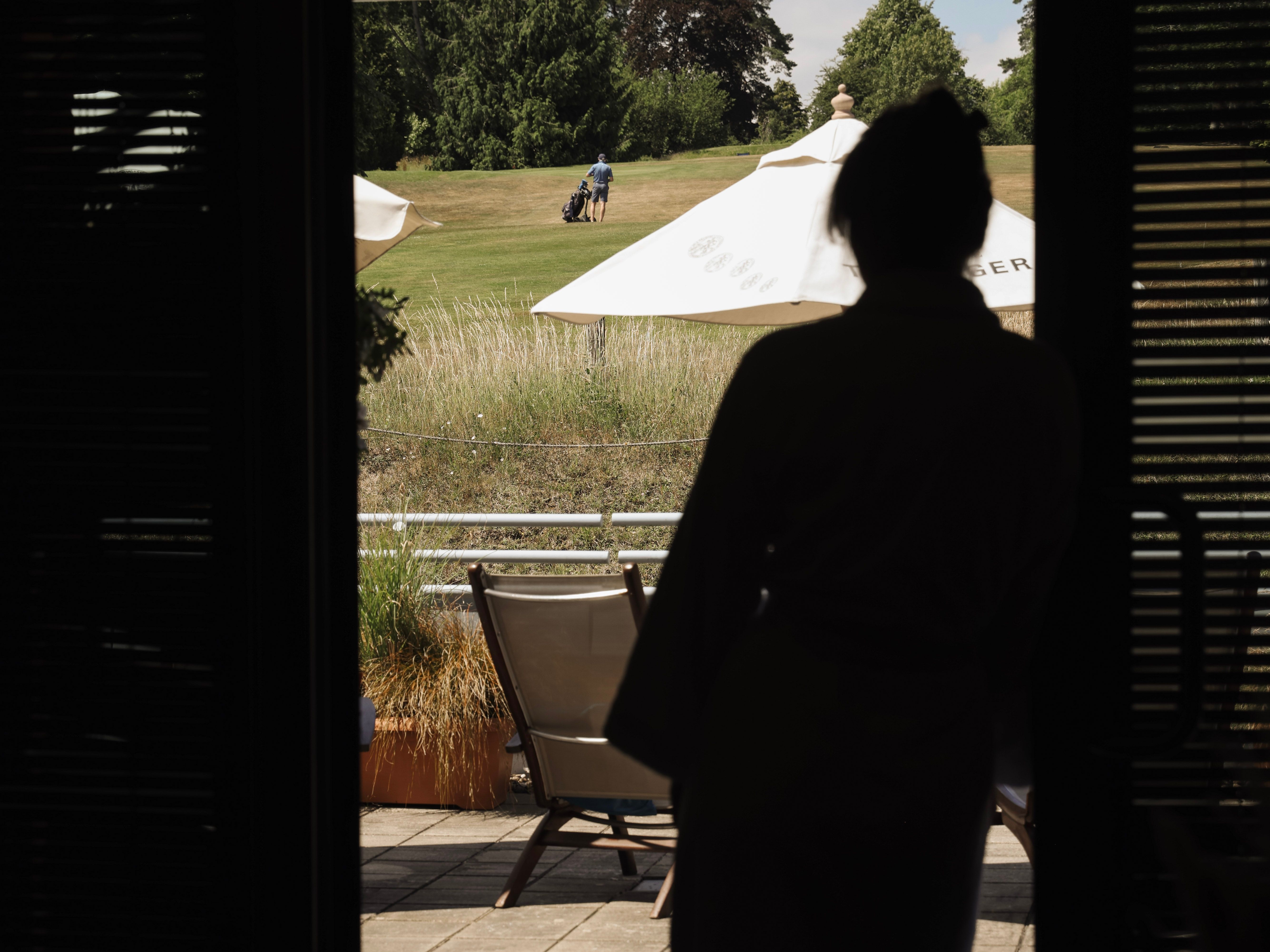 Silhouette of a person standing in a doorway looking outside at a person walking on a grassy field with a golf bag, with patio chairs and large umbrellas in the foreground.