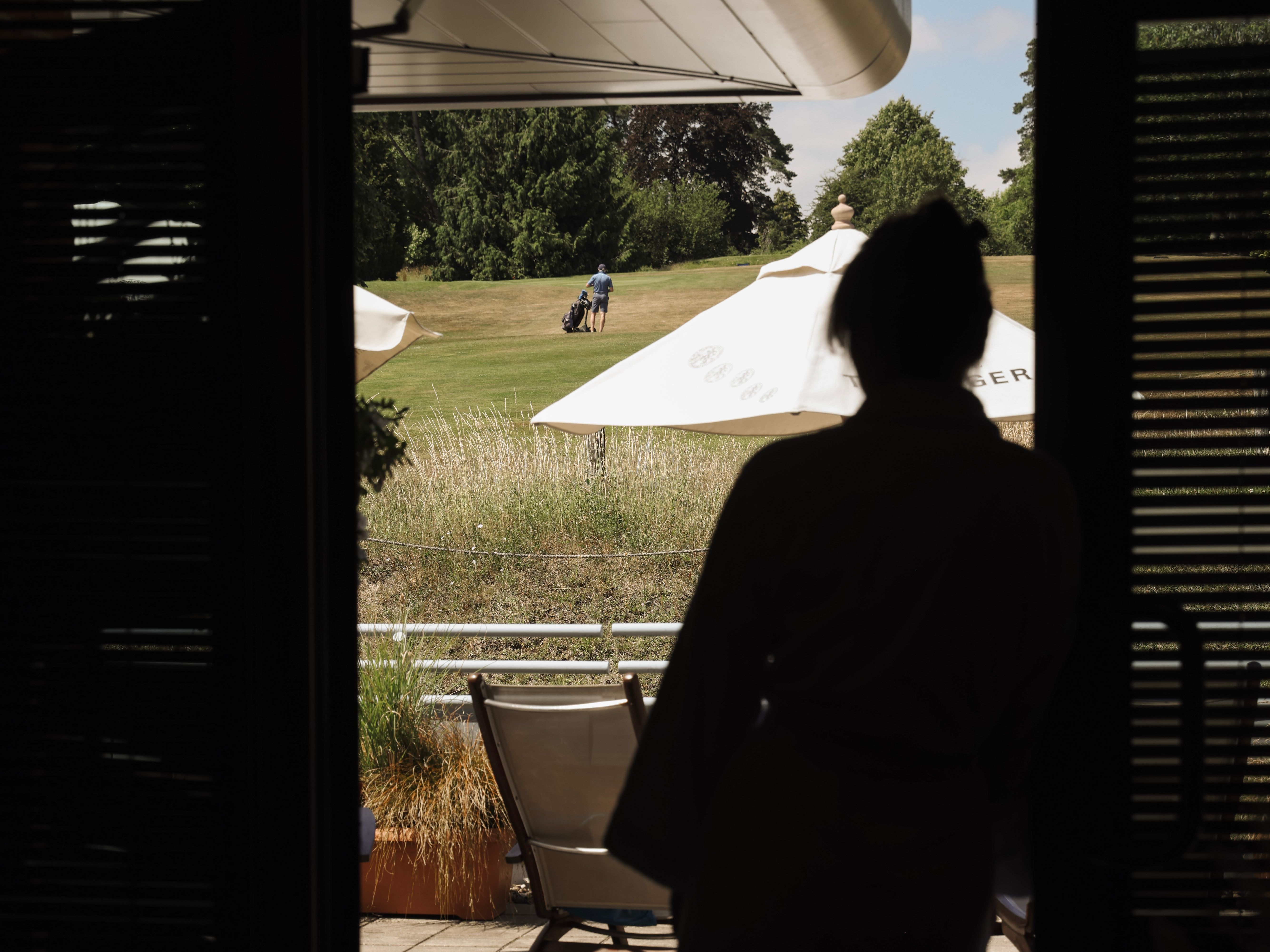 Silhouette of a person standing in a doorway looking outside at a person walking on a grassy field with a golf bag, with patio chairs and large umbrellas in the foreground.