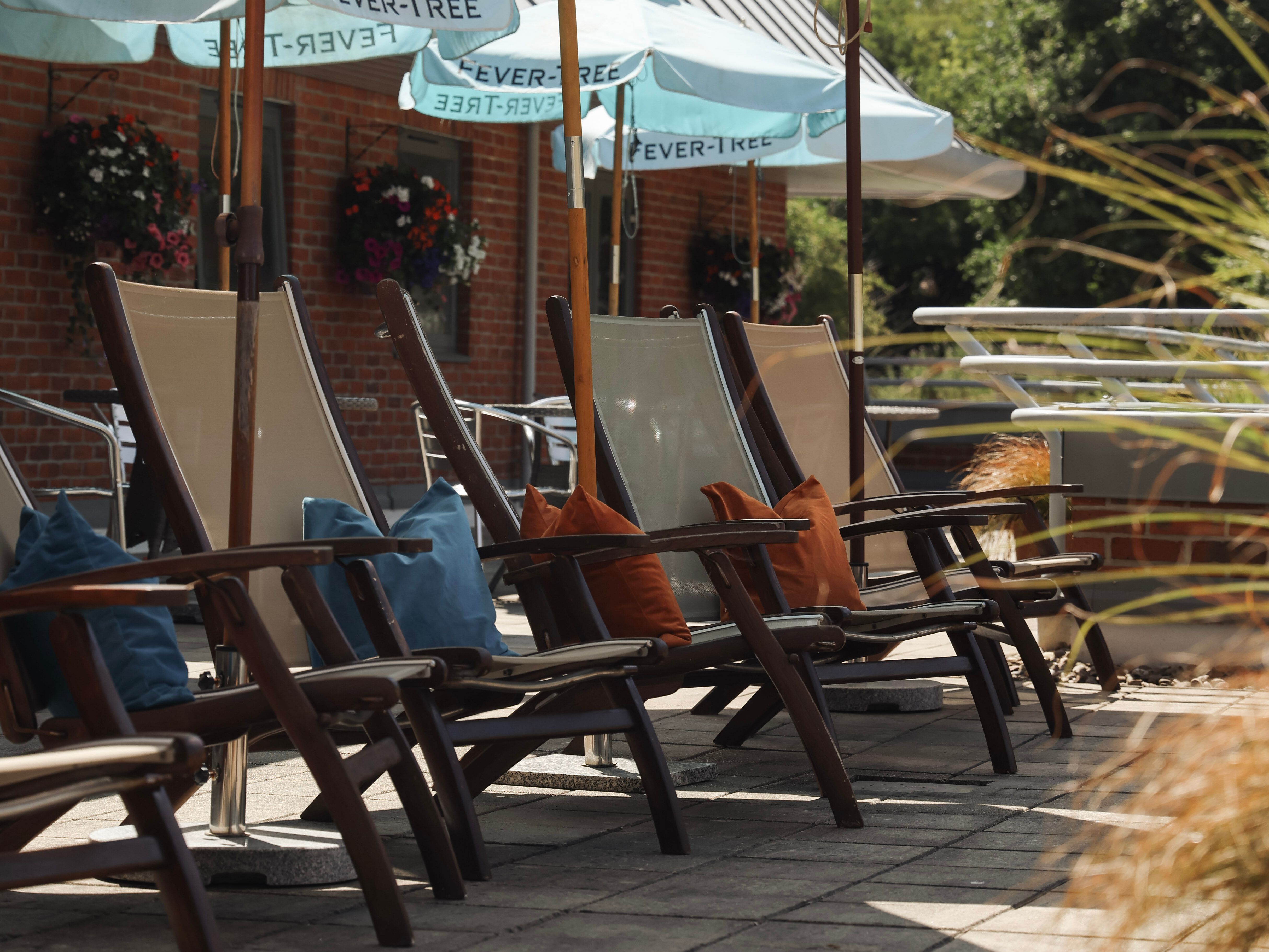 Outdoor wooden lounge chairs with colorful pillows under blue umbrellas at a patio area.