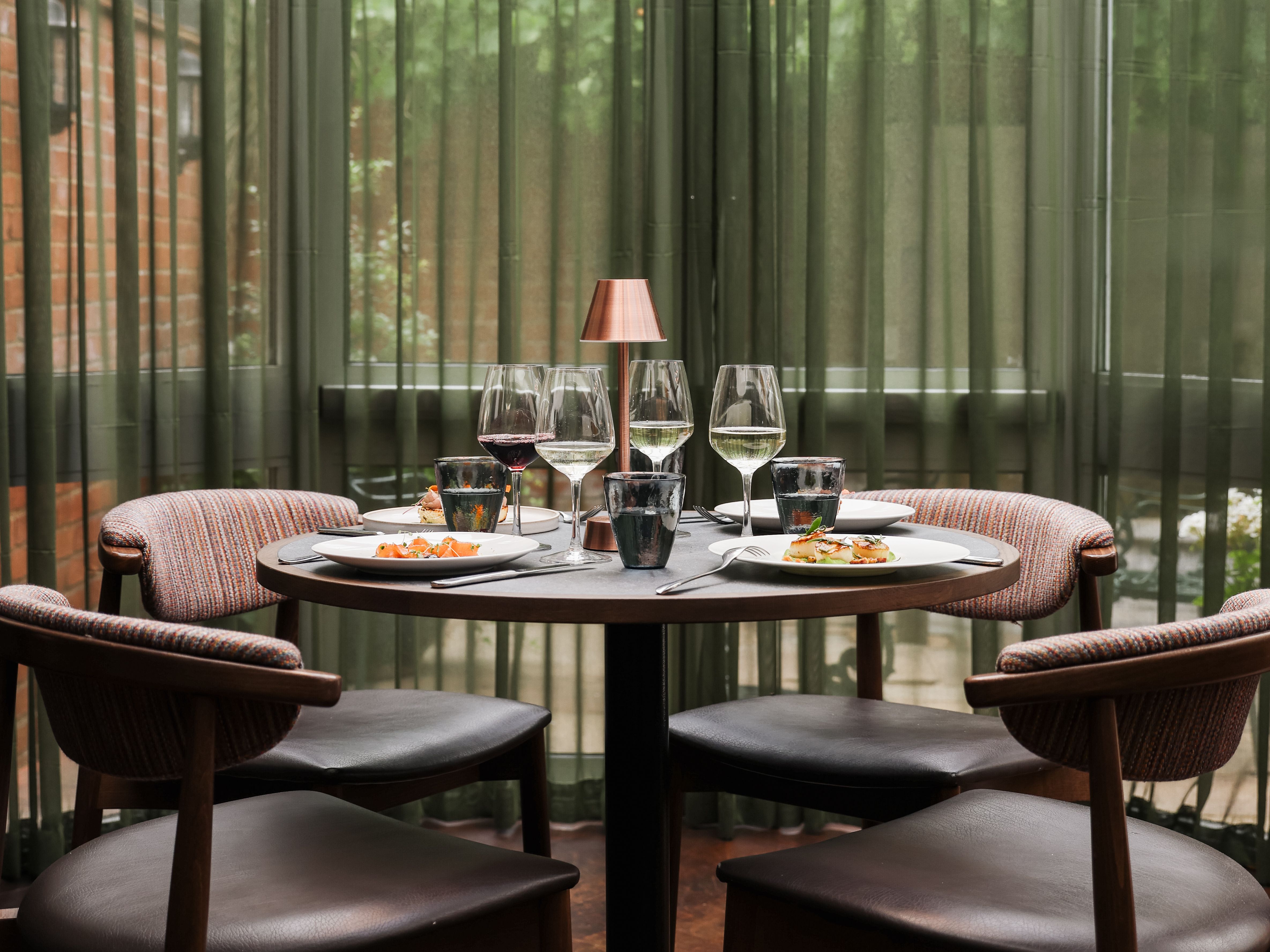 Elegant dining table set for four with wine glasses, plates of food, and a small lamp in a modern restaurant setting.